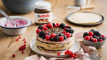 A Nutella® crepe cake, decorated with fresh berries is displayed on a plate. A jar of Nutella®, fresh berries and cream are shown in the background.
