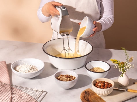 A baker holds a whisk in a large bowl and pours batter into it. Small bowls of brownie ingredients surround the bowl.