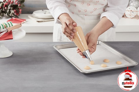 A baker is piping small dots of choux pastry onto a baking tray lined with parchment paper using a piping bag.