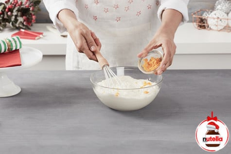 A baker is whisking orange peel with fresh cream in a large glass mixing bowl using a hand whisk.