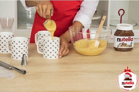 A baker is pouring mug cake mixture from a glass mixing bowl into 4 separate mugs.