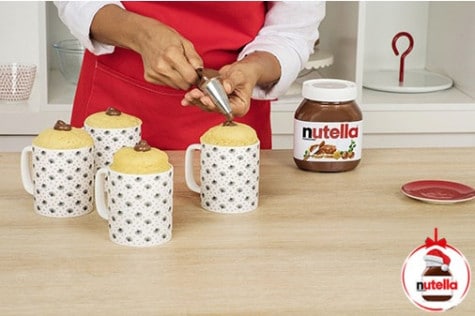 A baker is using a piping bag to fill each hole in the mug cakes with Nutella®.
