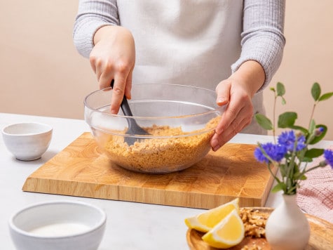 A baker is mixing chopped walnuts, cookie crumbs and butter in a glass mixing bowl using a silicone spatula.