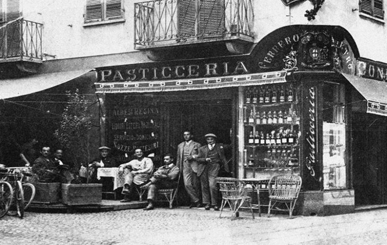 Vintage black and white photo of a "Pasticceria" shopfront with people gathered outside, some sitting, some standing